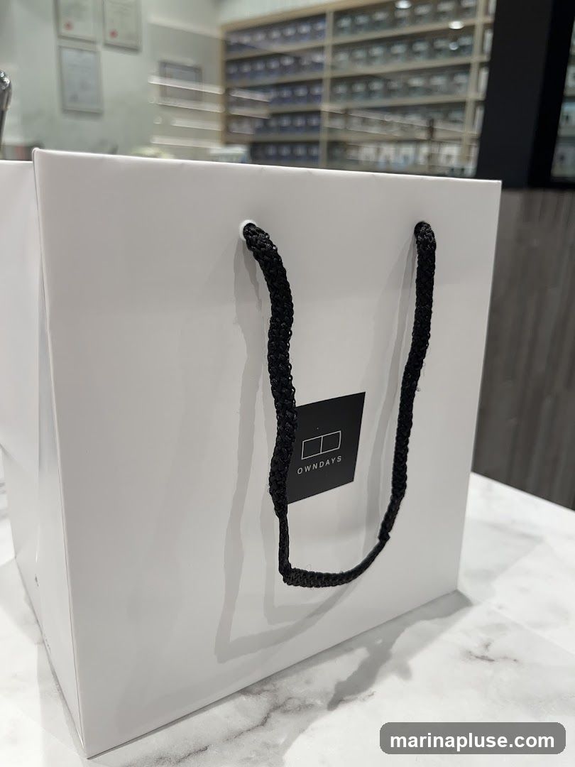 A white shopping bag with black rope handles sits on a marble-patterned surface. A black square logo with the text "OWNDAYS" is visible on the front of the bag. In the background, shelves filled with glasses are out of focus.