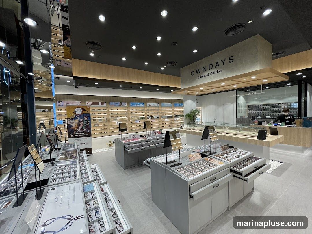 The interior of an eyewear store, featuring numerous displays of eyeglasses. In the foreground, tiered displays showcase various frames. In the midground, a large banner for a "Demon Slayer Frame Collection" is visible. Further back, a counter with a cashier and a wall lined with more eyewear options are present. The store has a modern design with a black ceiling and wooden accents.