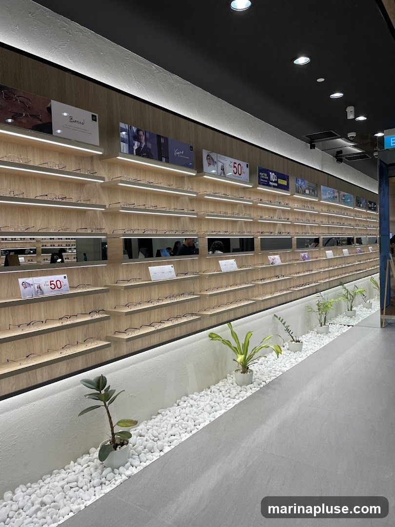A well-lit interior of an eyewear store featuring numerous shelves displaying a wide selection of eyeglasses. The shelves are organized and illuminated with LED lighting. Above the shelves, there are various promotional displays and advertisements, including one highlighting a "50% OFF" sale. The store has a modern aesthetic with wooden accents and a clean, minimalist design. Potted plants and white pebbles are used as decorative elements along the lower wall, adding a touch of nature to the space. The floor is a smooth, grey material.
