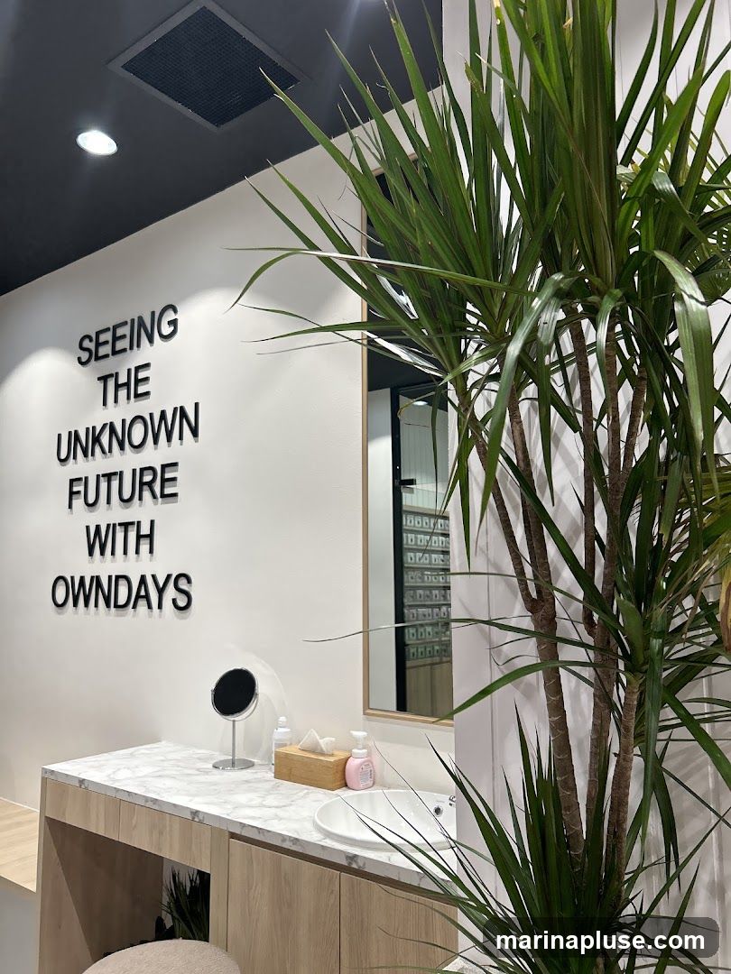 The image shows the interior of a store with a white wall featuring black text that reads "SEEING THE UNKNOWN FUTURE WITH OWNDAYS". To the right, there is a vanity area with a marble countertop, a sink, a mirror, a tissue box, and a bottle of soap. A large potted plant with long green leaves is prominently displayed in the foreground, partially obscuring the view of the vanity and a reflection in the mirror which shows shelves of products. The ceiling is black with recessed lighting.