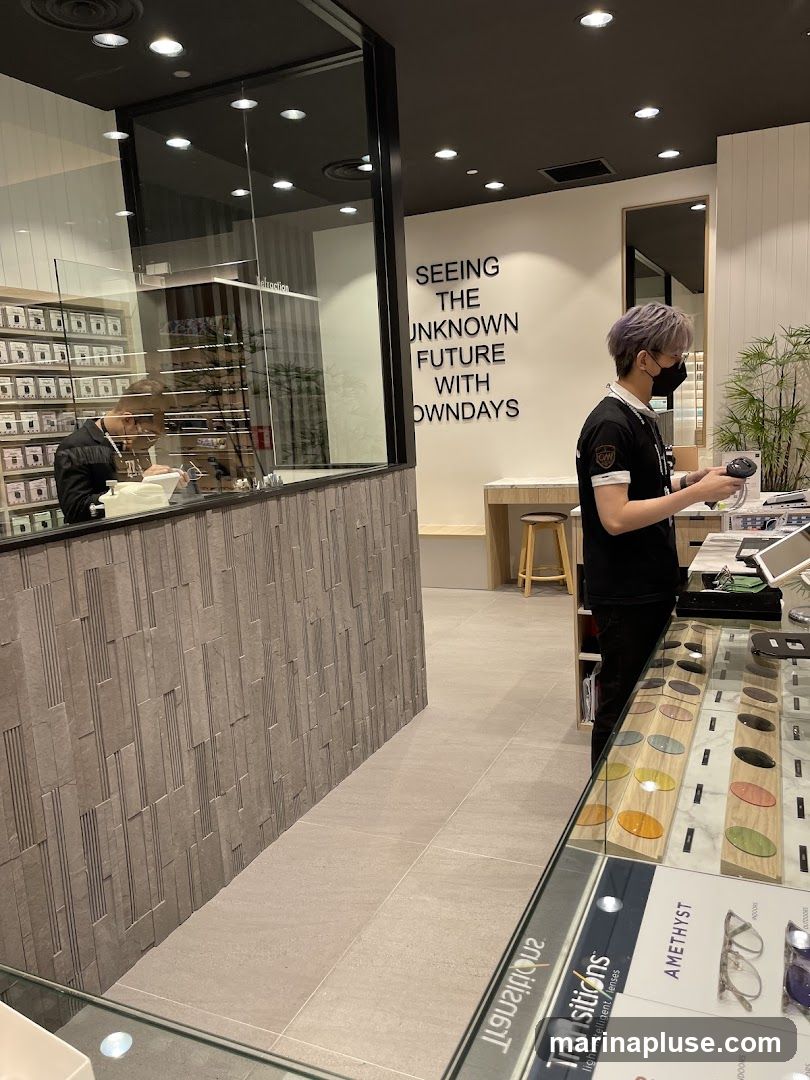 An interior view of an eyewear store. A person wearing a mask and a black shirt with a logo is standing behind a counter, holding a scanner. Display cases in the foreground show various lenses and eyeglass frames. On the wall, text reads "SEEING THE UNKNOWN FUTURE WITH OWNDAYS". In the background, through a glass partition, another person is seated at a desk, seemingly examining eyeglasses. Shelves filled with small boxes are visible on the left.