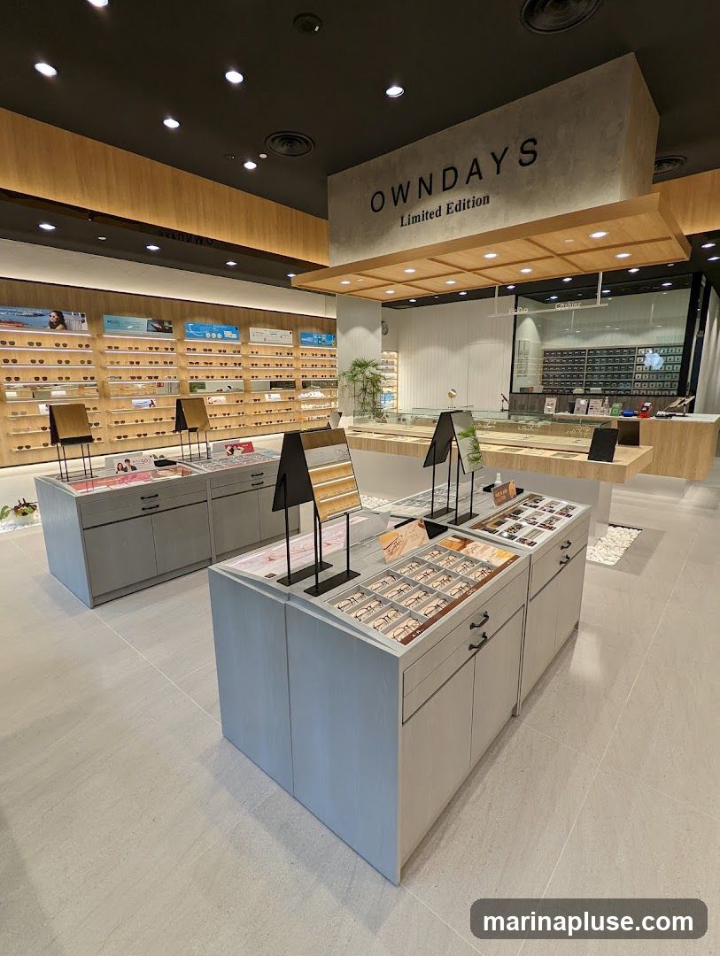 The interior of an eyewear store with displays of glasses. The store features a modern design with wooden accents and bright lighting. The brand name "OWNDAYS Limited Edition" is visible on a prominent sign. Shelves filled with various eyeglasses are displayed along the walls, and there are several display stands in the center of the store showcasing more eyewear.