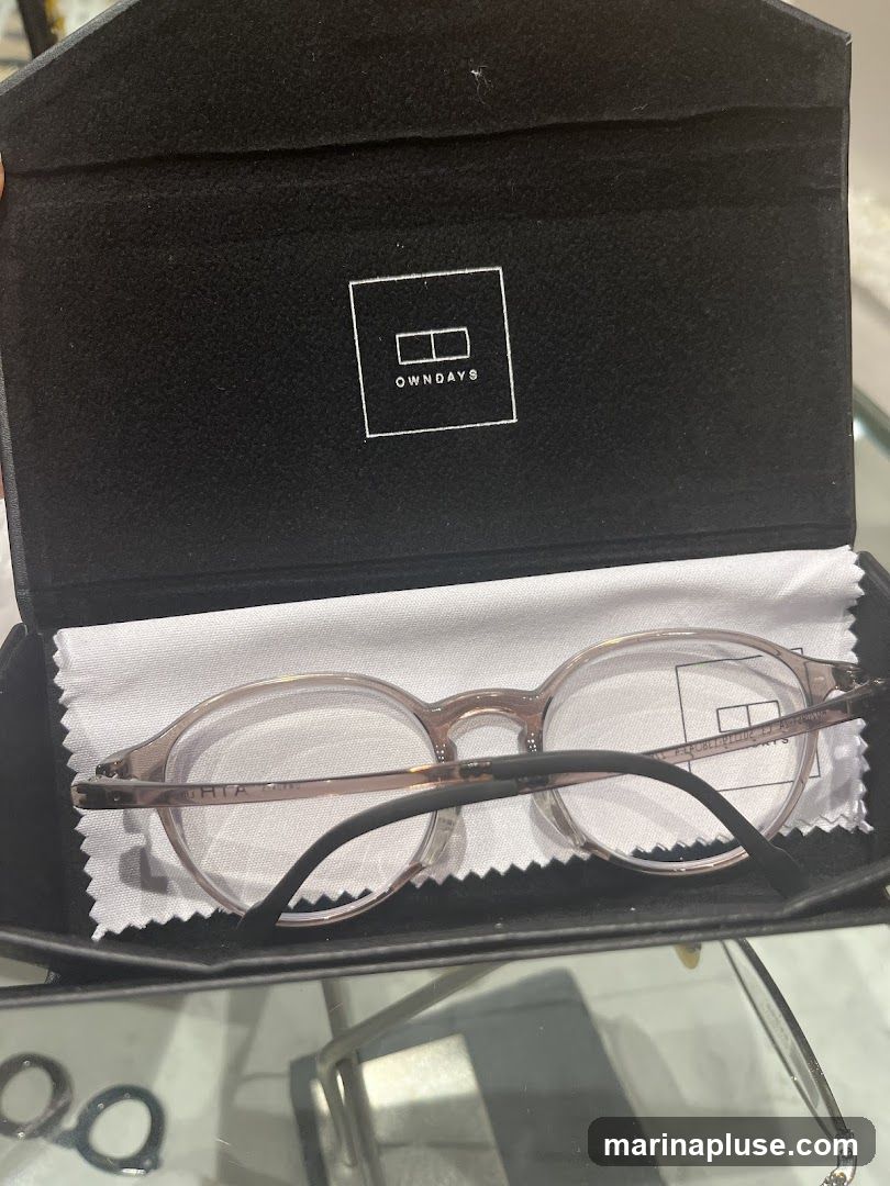 A pair of light pink eyeglasses with clear lenses are displayed inside a black case. The inside lid of the case has the OWNDAYS logo printed in white. A white cleaning cloth is placed under the eyeglasses.