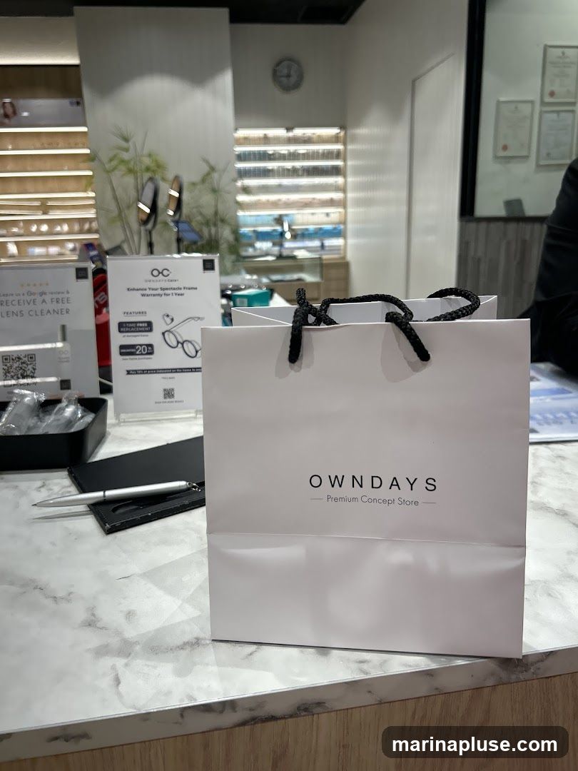 A white shopping bag with the OWNDAYS logo and "Premium Concept Store" printed on it sits on a marble counter. To the left, there are promotional signs, one offering a free lens cleaner for a Google review, and another detailing spectacle frame warranty. In the background, shelves display various eyeglasses and accessories.