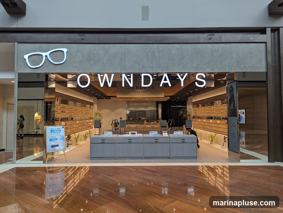 The exterior of an OWNDAYS optical shop. The shop has a large sign with the OWNDAYS logo and name. Inside, shelves are stocked with eyeglasses. There are also display stands and a promotional poster.