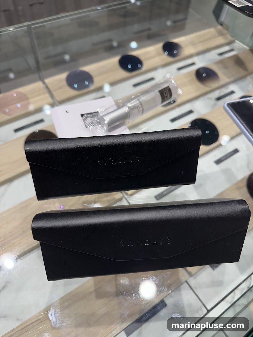 Two black eyeglass cases are displayed on a glass surface. The cases have the embossed text "OAKDAYS" on them. In the background, various lenses and a bottle of lens cleaner are visible on wooden stands within a display case.