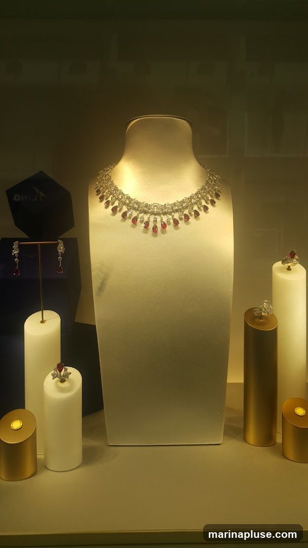 A display case featuring a diamond and ruby necklace on a mannequin bust. To the left, a pair of diamond and ruby earrings are displayed on a stand. To the right, two diamond rings are showcased on cylindrical pedestals. A dark blue hexagonal box with the word "CHAUMET" visible is also present. The display is illuminated with warm lighting.