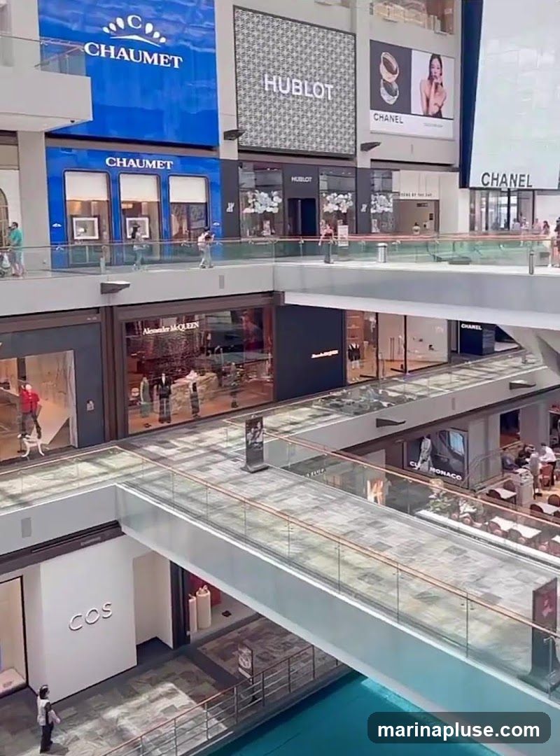 The image shows the interior of a modern shopping mall with multiple levels and walkways. Several high-end retail stores are visible, including Chaumet, Hublot, Chanel, Alexander McQueen, and COS. Large digital billboards display brand logos and advertisements. People are seen walking on the elevated walkways and inside the stores. The mall features a bright, open design with glass railings and a blue-tiled water feature visible at the bottom.