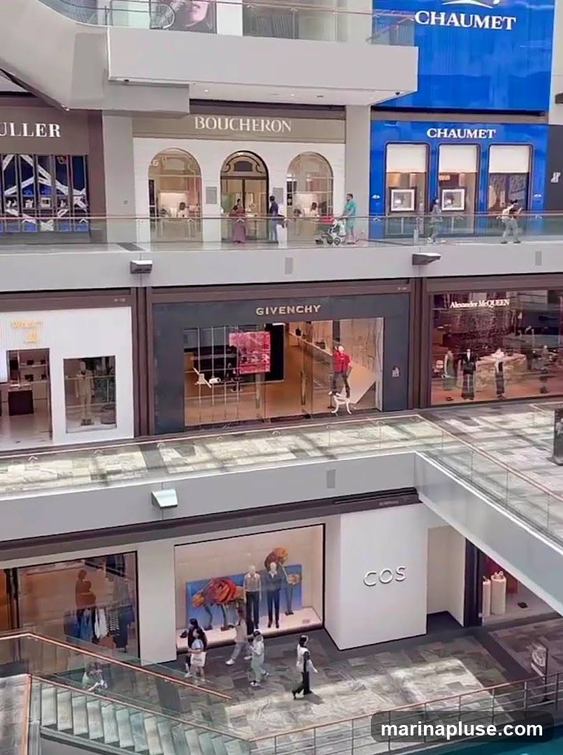 The image shows an interior view of a modern shopping mall with multiple levels. Several high-end retail stores are visible, including Boucheron, Chaumet, Givenchy, Alexander McQueen, and COS. People are seen walking on the elevated walkways and browsing in front of the shops. The architecture features clean lines, glass railings, and well-lit spaces.
