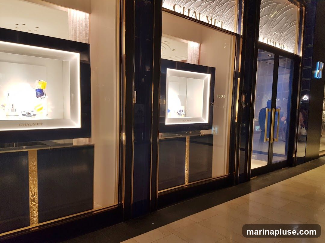 The exterior of a luxury jewelry and watch store. The storefront features large display windows showcasing watches and jewelry. The brand name "CHAUMET" is visible above the windows and on the display cases. The entrance has double glass doors with gold handles.