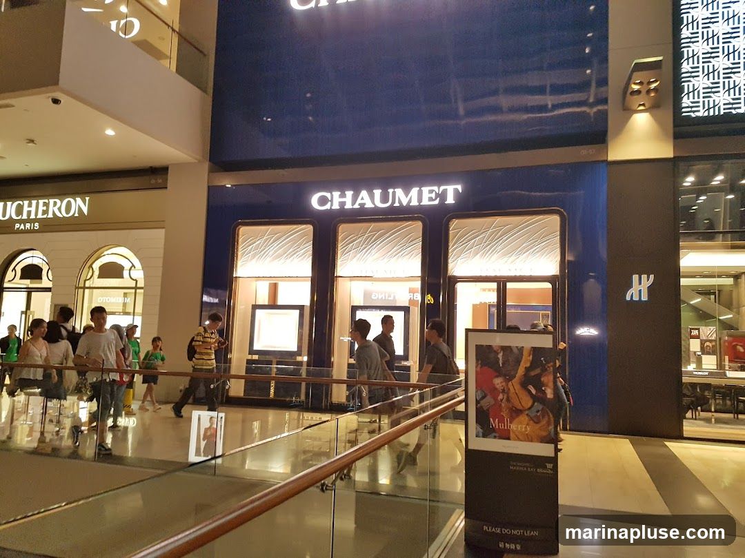 A wide shot of a shopping mall interior, showcasing the storefronts of luxury brands. On the right, the prominent blue facade of CHAUMET is visible, with its name displayed in large white letters. Below the name, three illuminated display windows showcase jewelry or watches. To the left of CHAUMET, another store with a dark facade features a blue logo. Further to the left, the store VACHERON CONSTANTIN PARIS is visible with its name above arched entrances. Several shoppers are walking in the mall. A display stand with an advertisement for Mulberry is positioned in the foreground.