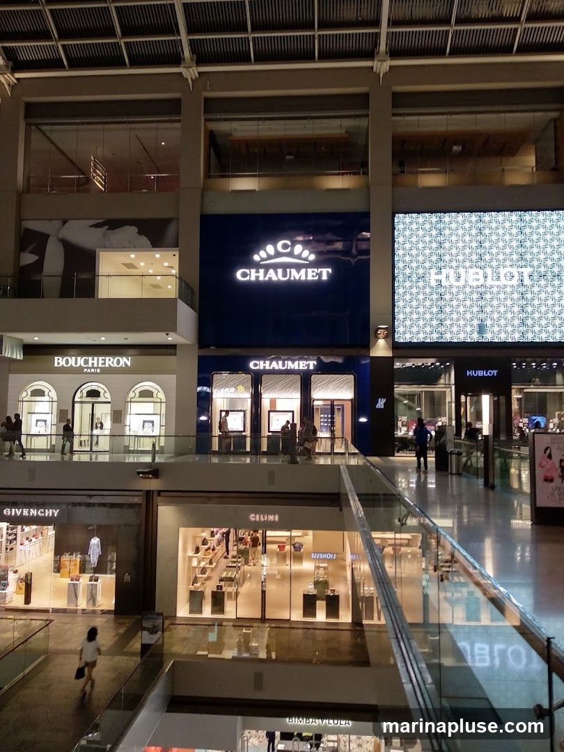 A view of several high-end retail stores within a modern shopping mall. The image captures the storefronts of Boucheron, Chaumet, Hublot, Givenchy, and Celine, among others. The stores feature illuminated displays showcasing merchandise, and shoppers can be seen browsing inside. The overall ambiance is one of luxury and sophistication, with polished floors and well-lit interiors.