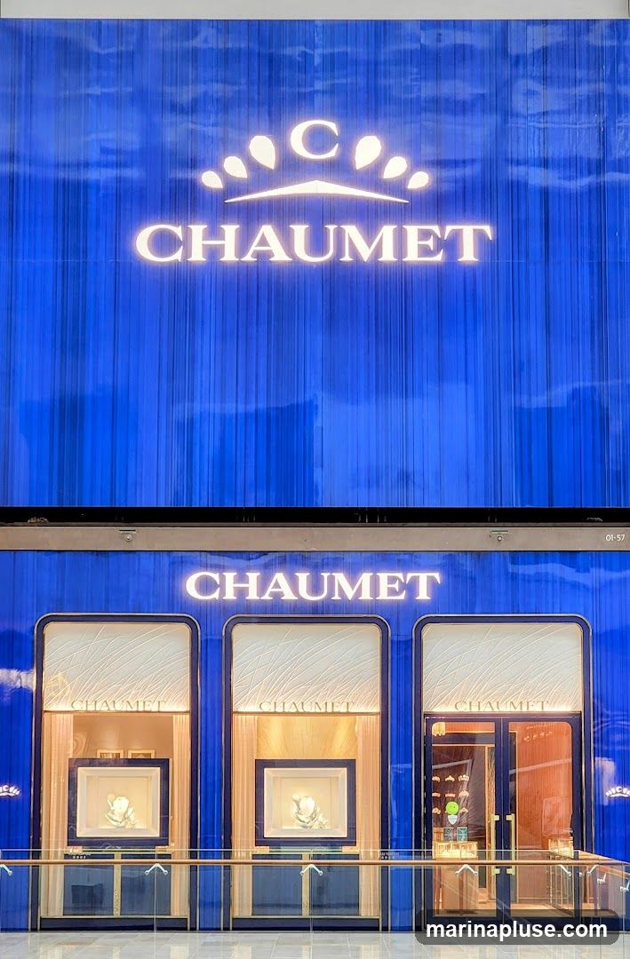 The exterior of a luxury jewelry store, Chaumet, with a prominent illuminated logo and name displayed on a blue, curtain-like facade. Below the main signage, there are three display windows showcasing jewelry. The windows are framed in dark blue and gold, with soft lighting illuminating the displayed pieces. The rightmost window appears to be an entrance with glass doors.