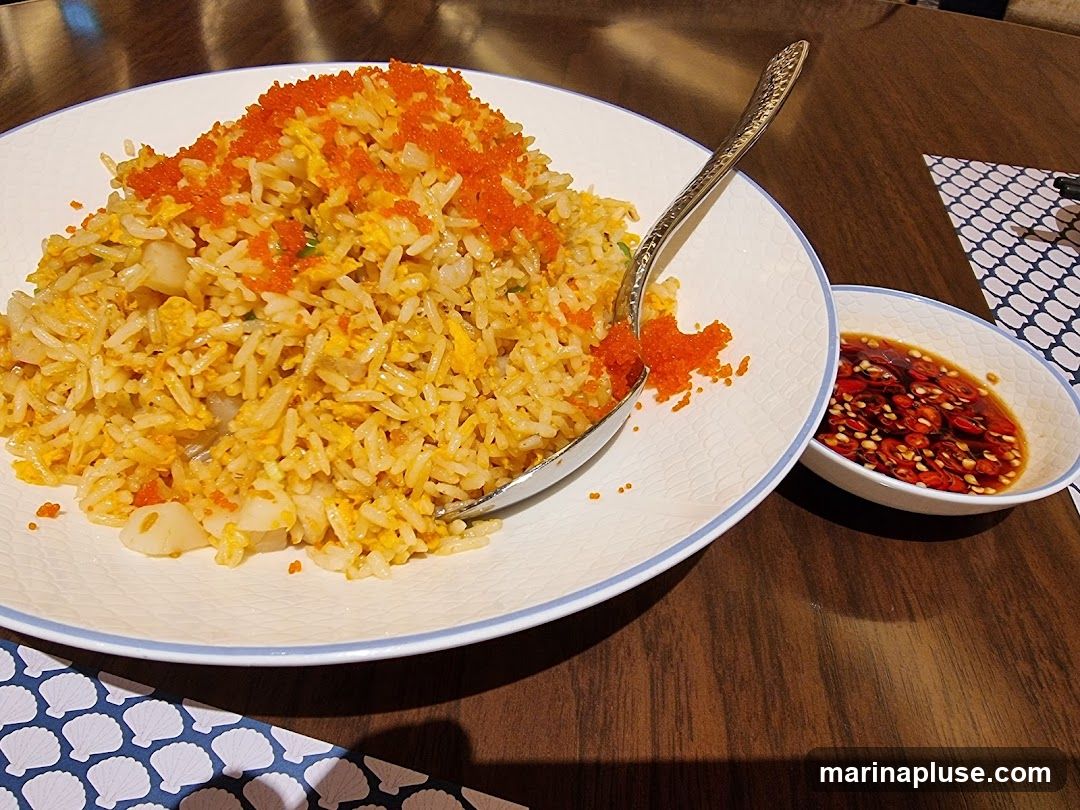 A plate of seafood fried rice topped with tobiko (flying fish roe) is served with a small bowl of chili dipping sauce. The fried rice contains visible pieces of seafood, likely scallops, and scrambled egg. A spoon rests on the plate. The meal is presented on a wooden table with a patterned placemat.