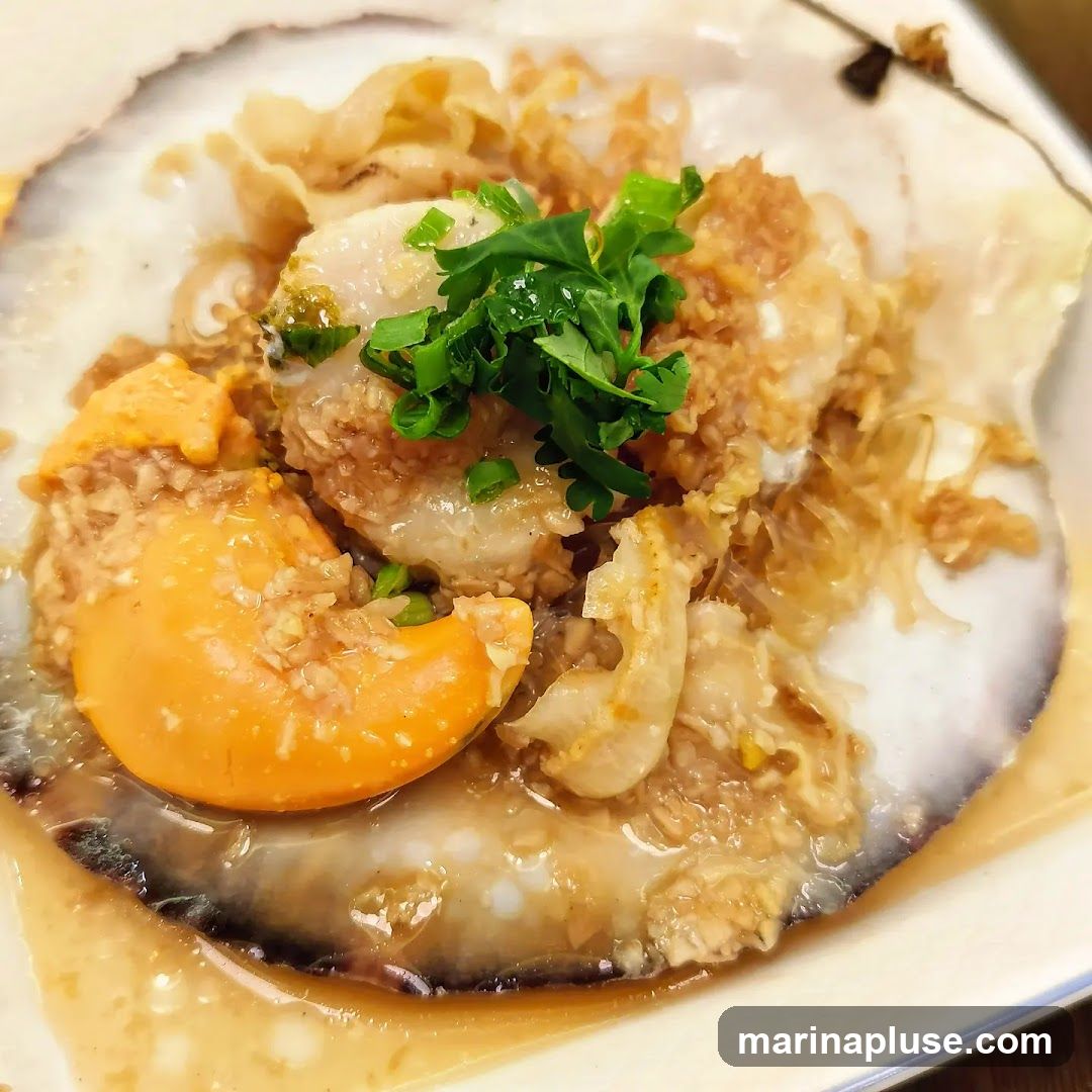 A close-up shot of a steamed scallop served in its shell. The scallop is topped with minced garlic, crispy fried shallots, and garnished with fresh green herbs. A bright orange egg yolk is visible on the side, and the dish is served in a light sauce with glass noodles underneath.