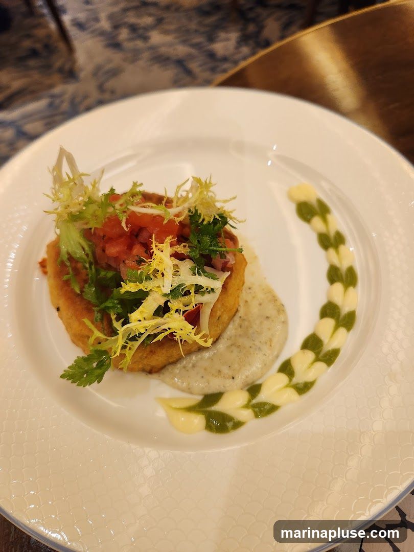 A close-up shot of a crab cake served on a white plate. The crab cake is topped with a fresh salsa and garnished with frilly lettuce and parsley. A dollop of creamy aioli and a decorative swirl of green sauce complete the presentation. The plate has a subtle embossed pattern around the rim.
