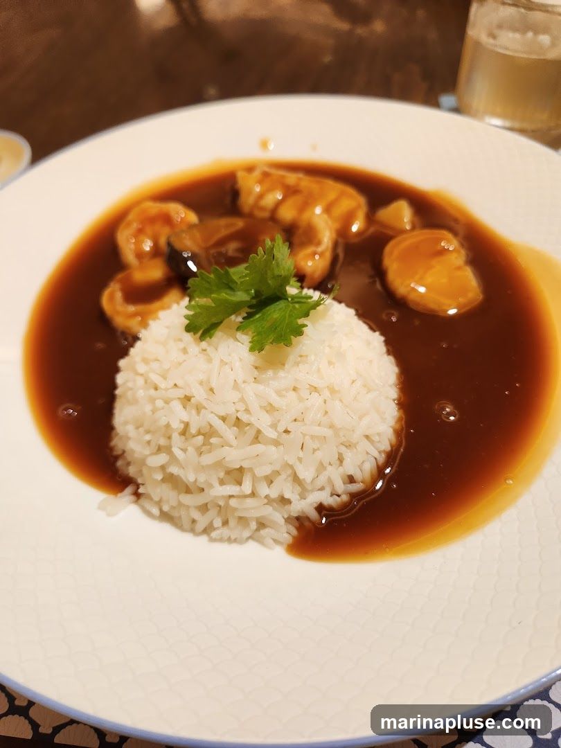 A close-up shot of a white plate with a mound of white rice. The rice is partially covered by a dark, glossy sauce containing pieces of braised food, possibly meat or seafood, and some vegetables. A sprig of fresh cilantro garnishes the dish. A glass of a light-colored beverage is visible in the blurred background.