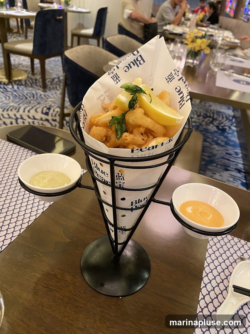A cone of fried calamari, garnished with lemon wedges and herbs, is presented in a restaurant setting. Two small bowls of dipping sauces, one creamy green and the other orange, are attached to the cone holder. The table is set with blue and white patterned placemats. In the background, diners are seated at tables with blue chairs and elegant decor.