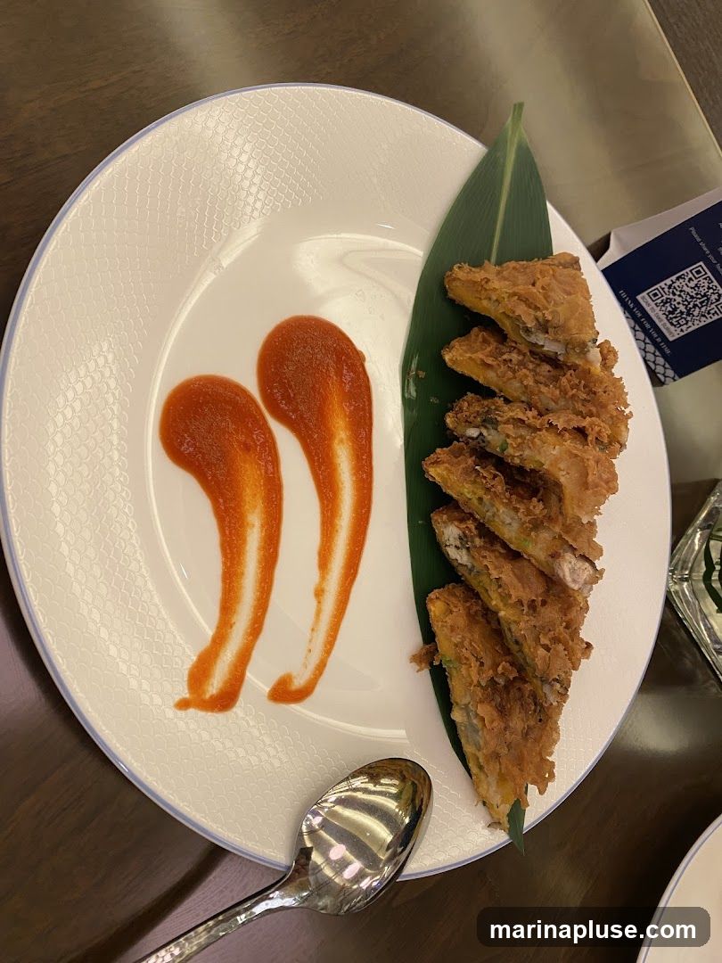 A white plate with a textured rim holds several pieces of fried food, arranged on a green leaf. Two dollops of red sauce are on the left side of the plate. A spoon rests on the table in front of the plate. A QR code on a small card is visible in the upper right corner.