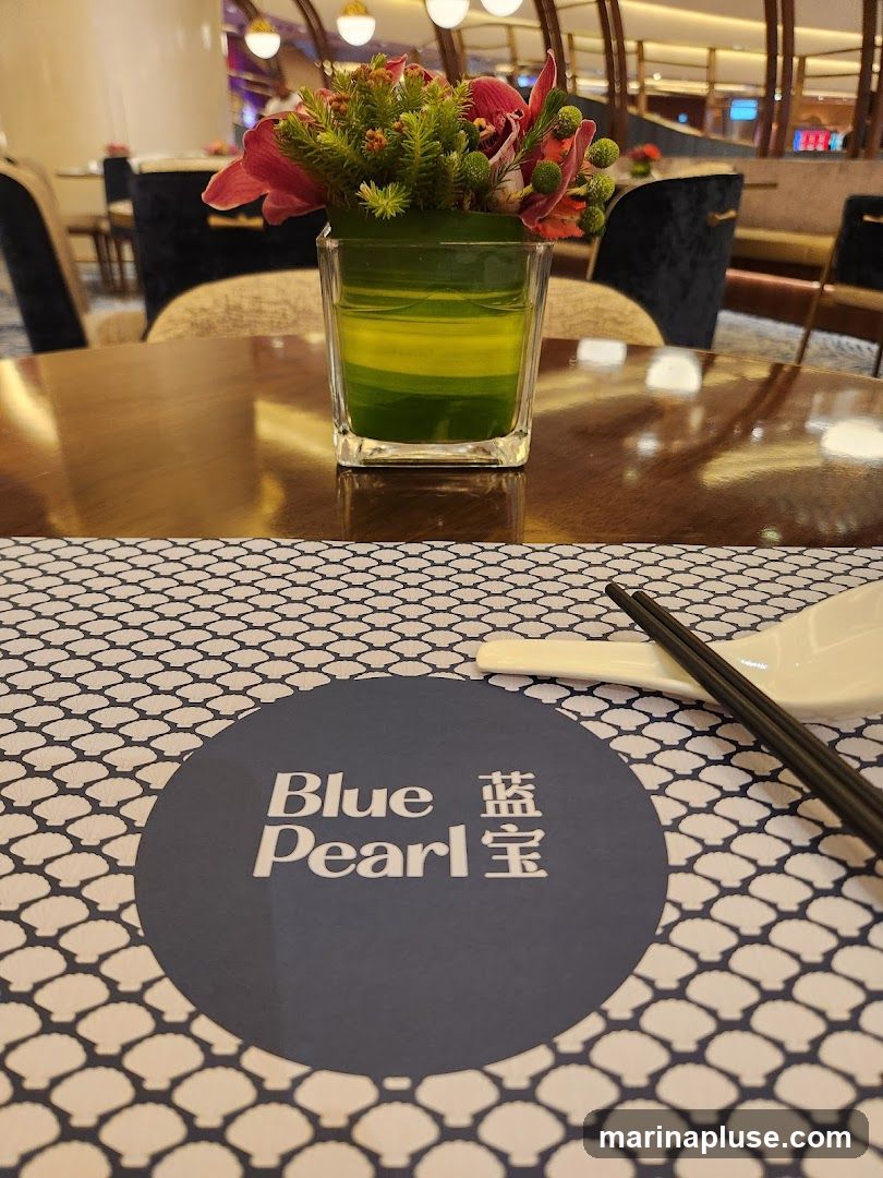 A close-up view of a table setting in a restaurant. A floral arrangement in a glass vase sits on a polished wooden table. A placemat with a seashell pattern is visible, featuring a dark blue circular logo with the text "Blue Pearl" and Chinese characters. Chopsticks and a spoon are placed on the placemat. The background shows the interior of the restaurant with seating and decorative lighting.