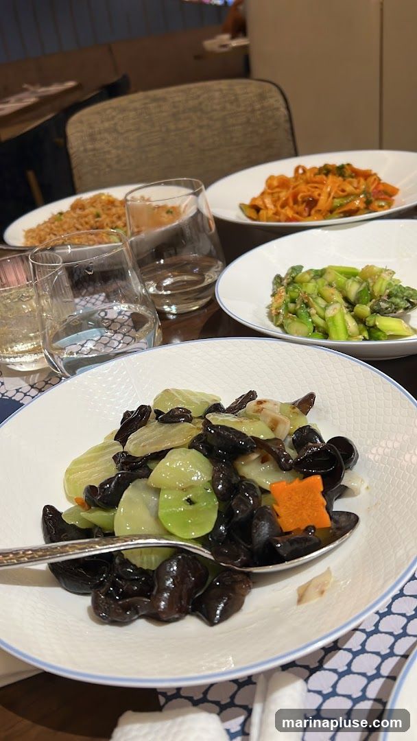 A table setting with several plates of food and glasses of drinks. In the foreground, a white plate holds a stir-fry dish with dark mushrooms, green vegetables, and carrot pieces. A spoon rests on the plate. In the background, other plates contain fried rice and a pasta dish with red sauce. Two glasses filled with a clear liquid are also visible. The setting appears to be in a restaurant with upholstered chairs and tables.