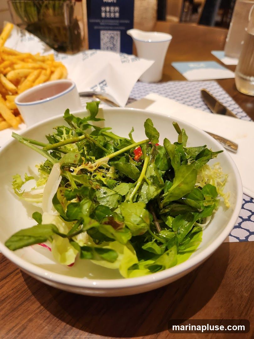 A close-up shot of a salad in a white bowl, with a side of french fries and a small dish of sauce in the background. The salad appears to be a mix of greens, possibly with some radish. The fries are golden brown and piled high. There is also a white pitcher and a glass of water on the table. A blue and white patterned placemat is visible under the food. A QR code is visible on a blue sign in the background.