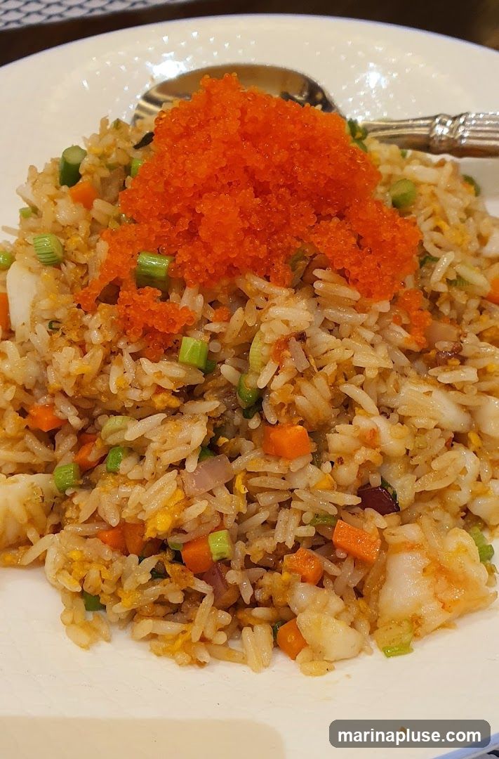 A close-up shot of a plate of fried rice topped with tobiko (flying fish roe). The fried rice contains visible pieces of scrambled egg, diced carrots, green onions, and what appears to be seafood. A spoon rests on the side of the plate.