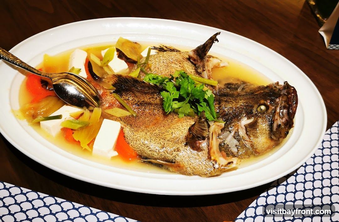 A whole steamed fish is served in a white oval dish with a light broth. The fish is garnished with fresh cilantro and surrounded by cubes of white tofu, sliced carrots, and leeks. A decorative spoon rests on the side of the dish. The dish is placed on a wooden table, with a patterned napkin visible in the foreground.