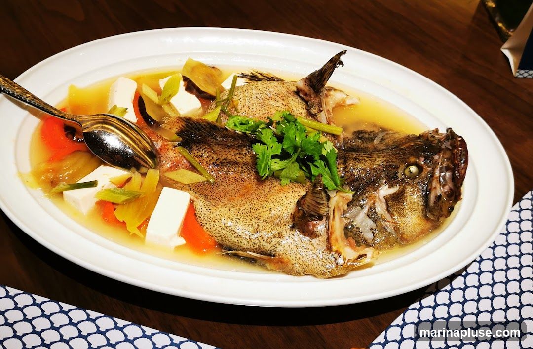 A whole steamed fish is served in a white oval dish with a light broth. The fish is garnished with fresh cilantro and surrounded by cubes of white tofu, sliced carrots, and leeks. A decorative spoon rests on the side of the dish. The dish is placed on a wooden table, with a patterned napkin visible in the foreground.