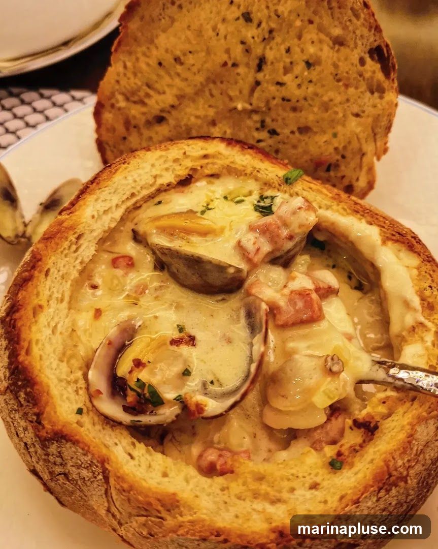 A close-up shot of a creamy clam chowder served in a hollowed-out bread bowl. The chowder is filled with clams, potatoes, and pieces of bacon, and is garnished with fresh parsley. A slice of crusty bread is placed behind the bowl.