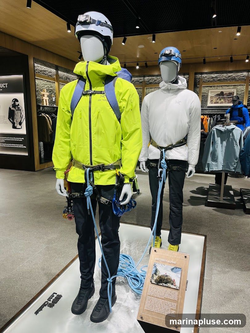 Two mannequins are displayed in a store, dressed in outdoor climbing gear. The mannequin on the left wears a bright yellow jacket, black pants, and a white helmet with a headlamp. It is equipped with a climbing harness, ropes, and carabiners. The mannequin on the right wears a white hooded jacket, black pants, and a blue helmet. Both mannequins are positioned on a display that resembles a rocky, snowy surface. In the background, racks of clothing and other merchandise are visible, suggesting an outdoor apparel store. A sign with an image and text is placed in front of the mannequin on the right.