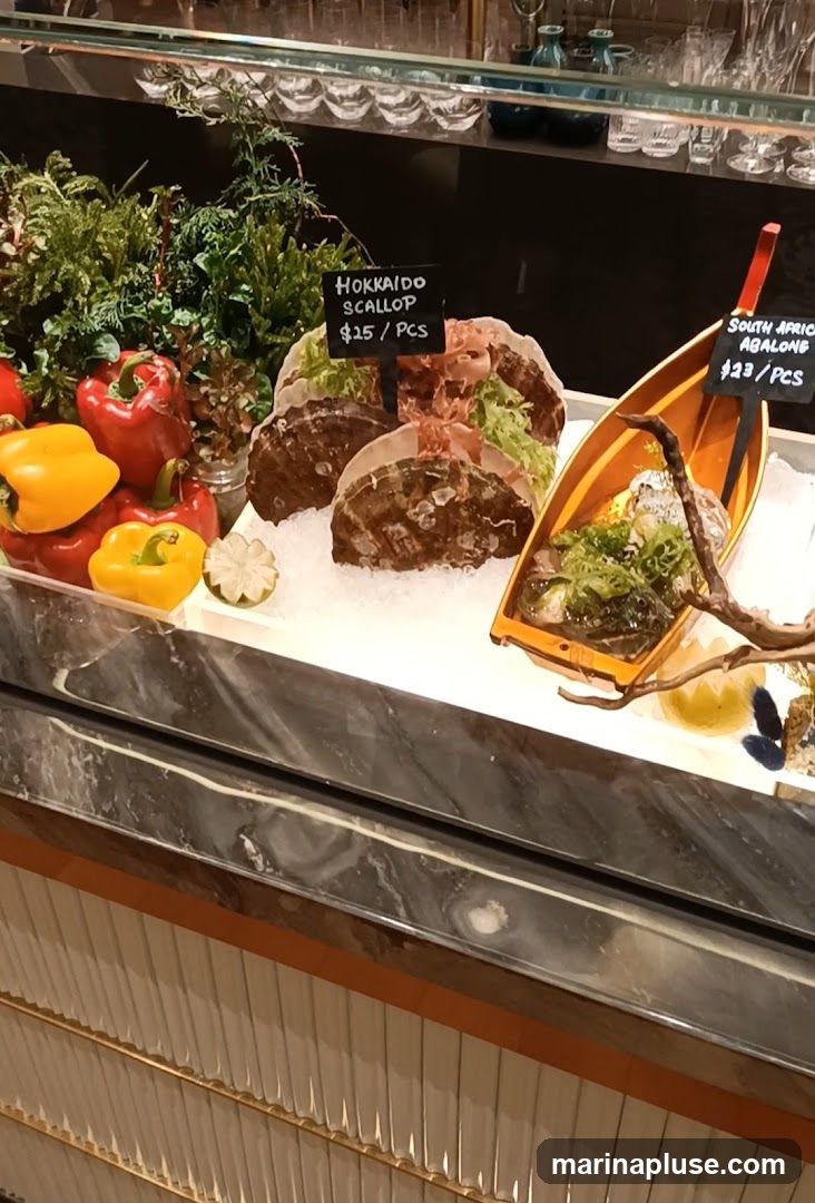 A display of fresh seafood on ice, including Hokkaido scallops and South African abalone. There are also bell peppers and leafy greens in the display. Small chalk signs indicate the prices: Hokkaido Scallop $25/pcs and South African Abalone $23/pcs.