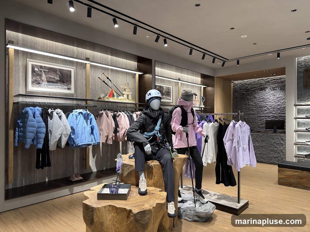 The interior of an outdoor apparel store features mannequins dressed in hiking gear, with racks of colorful jackets and pants displayed. Artwork and decorative elements related to outdoor activities are visible on the walls. The store has a modern and clean aesthetic with wooden flooring and stone accents.