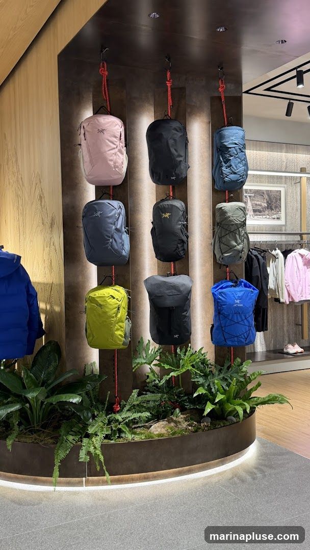 A retail display featuring nine backpacks of various colors, including pink, black, blue, grey, and lime green, suspended by red ropes against a dark textured wall. The display is accented with lush green plants in a curved planter at the base. In the background, clothing racks hold jackets and shirts, and a framed picture hangs on the wall.