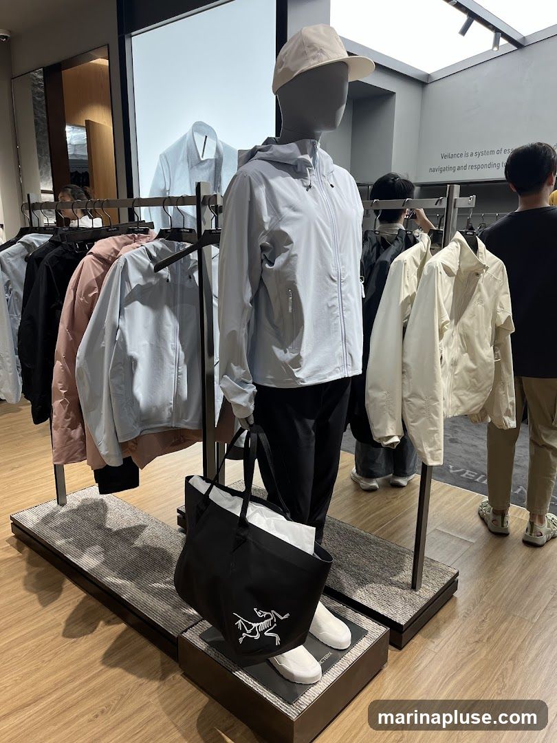 A mannequin wearing a light blue hooded jacket and a beige cap stands in a clothing store. The mannequin holds a black tote bag with a white logo. Racks of clothing, including jackets and shirts in various colors, are displayed around the mannequin. Several people are visible in the background, browsing the store. A sign on the wall reads "Veilance is a system of essential... navigating and responding to..."