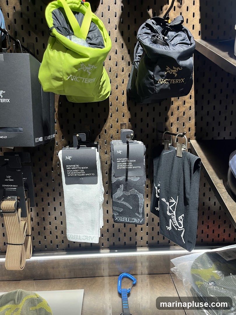 A display of outdoor gear, including two chalk bags (one lime green, one black), a pair of white socks, a pair of black camouflage socks, and a black garment with a white skeleton graphic. A tan conveyor belt is also visible. The items are displayed on a pegboard wall with the Arc'teryx logo present on several items.