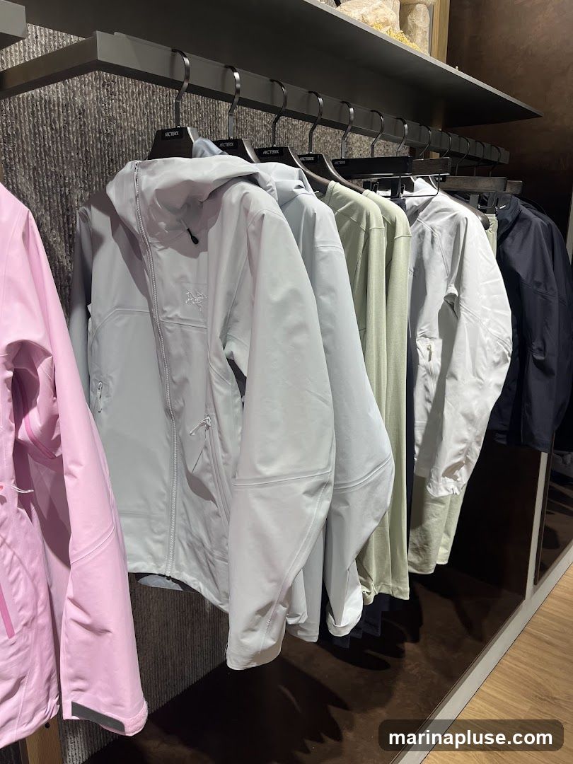 A rack of outdoor jackets in various colors, including pink, light gray, sage green, and black, are displayed on hangers in a store. The jackets appear to be made of technical fabric and feature zippers and hoods. The Arc'teryx logo is visible on some of the hangers.