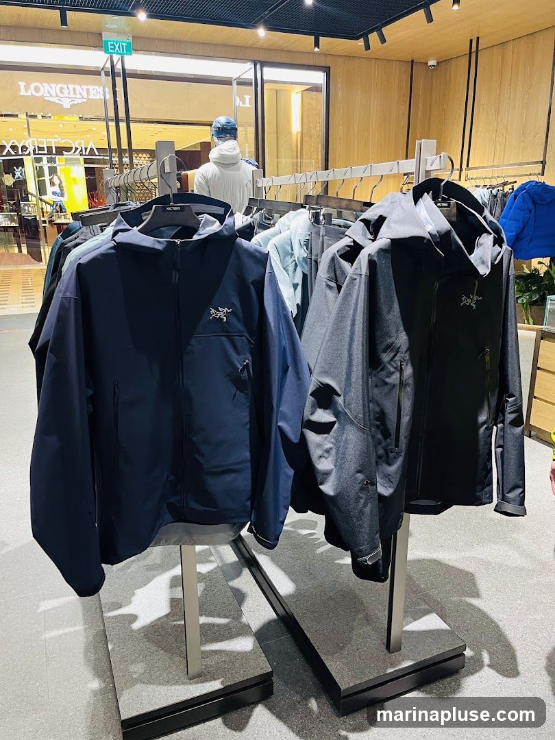 A retail display featuring several outdoor jackets on hangers. The jackets are primarily dark blue and black, with visible brand logos. In the background, a mannequin is dressed in a white hoodie and blue cap, and signage for 'LONGINES' and 'ARC' is visible through a glass partition, suggesting a high-end retail environment.