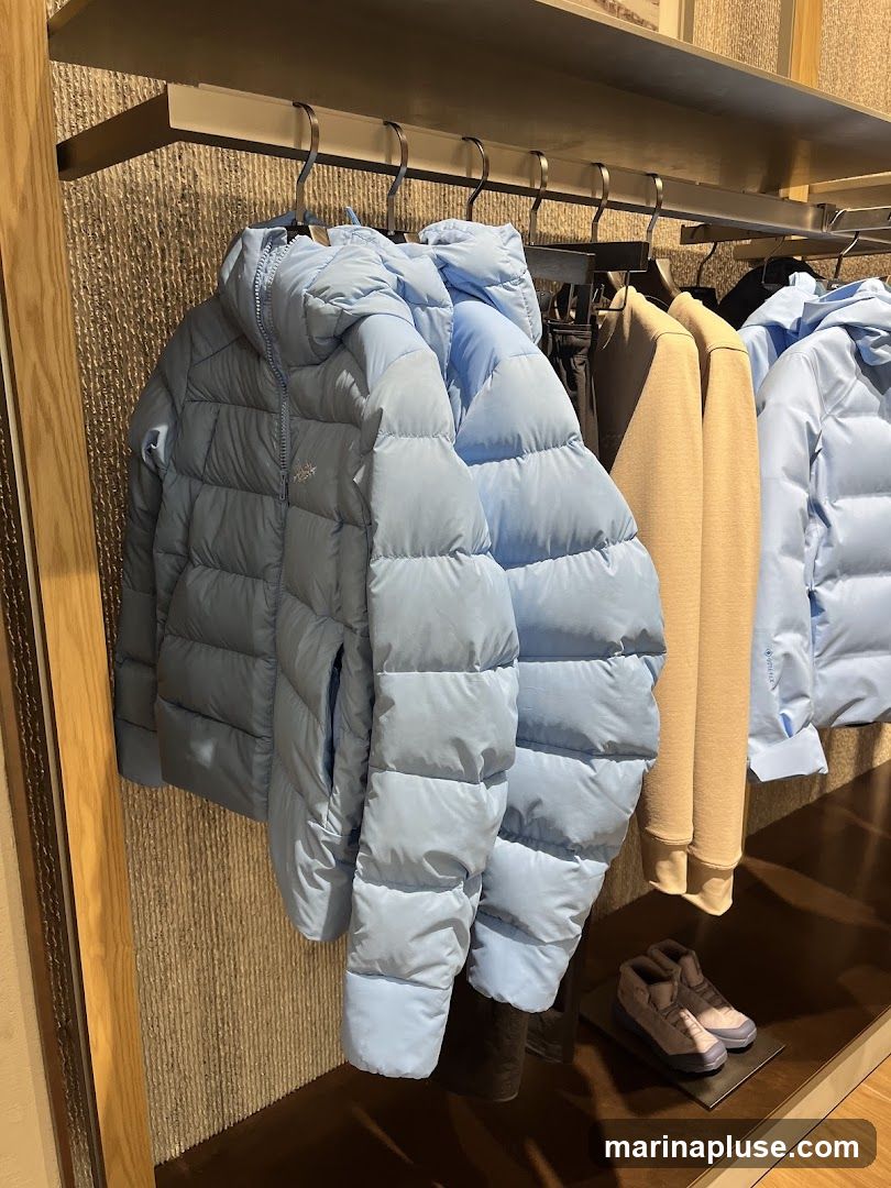 A display of light blue puffer jackets and a tan sweatshirt hanging on a rack in a store. A pair of grey and purple hiking boots are displayed on a shelf below. The background has a textured wall and a wooden frame.