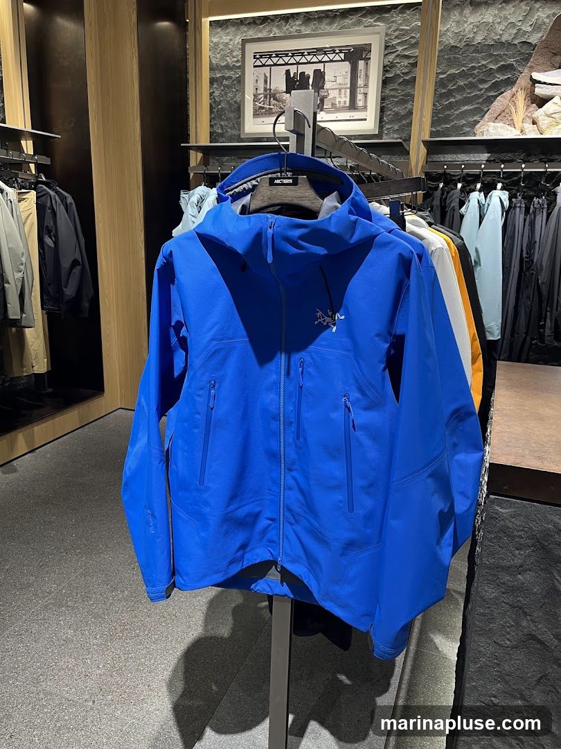 A bright blue jacket is displayed prominently on a hanger in what appears to be a retail store. The jacket has a hood and multiple zippered pockets. Behind it, other clothing items are visible on racks, including jackets and pants in various colors. A framed picture hangs on the wall in the background, and the store's interior features wooden accents and stone-like textured walls. The brand name 'ARC'TERYX' is visible on the hanger.