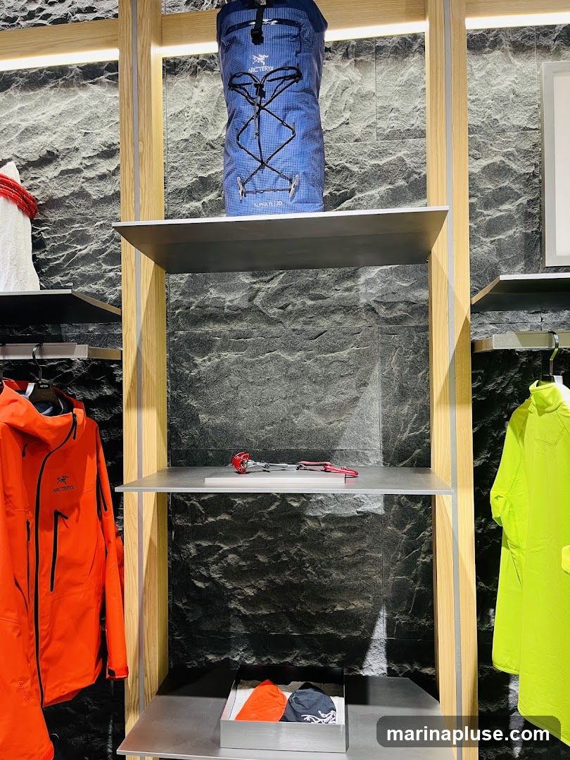 A display shelf featuring outdoor gear. On the top shelf, a blue Arc'teryx backpack is shown. The middle shelf has climbing equipment. The bottom shelf displays folded orange and dark blue accessories. To the left, an orange Arc'teryx jacket hangs, and to the right, a bright yellow jacket is displayed.