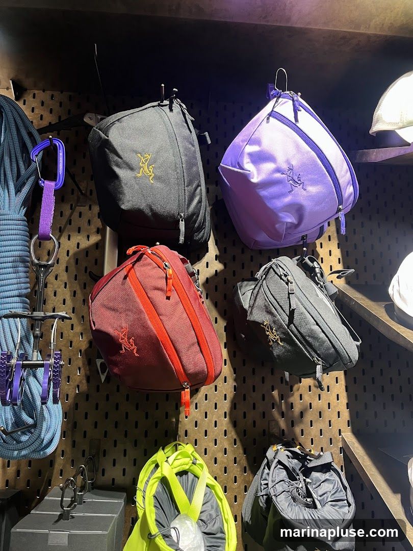 A wall display featuring various outdoor gear items, including backpacks, climbing ropes, and carabiners. Several bags are visible in different colors: black, purple, maroon, and lime green. Each bag has a prominent logo.