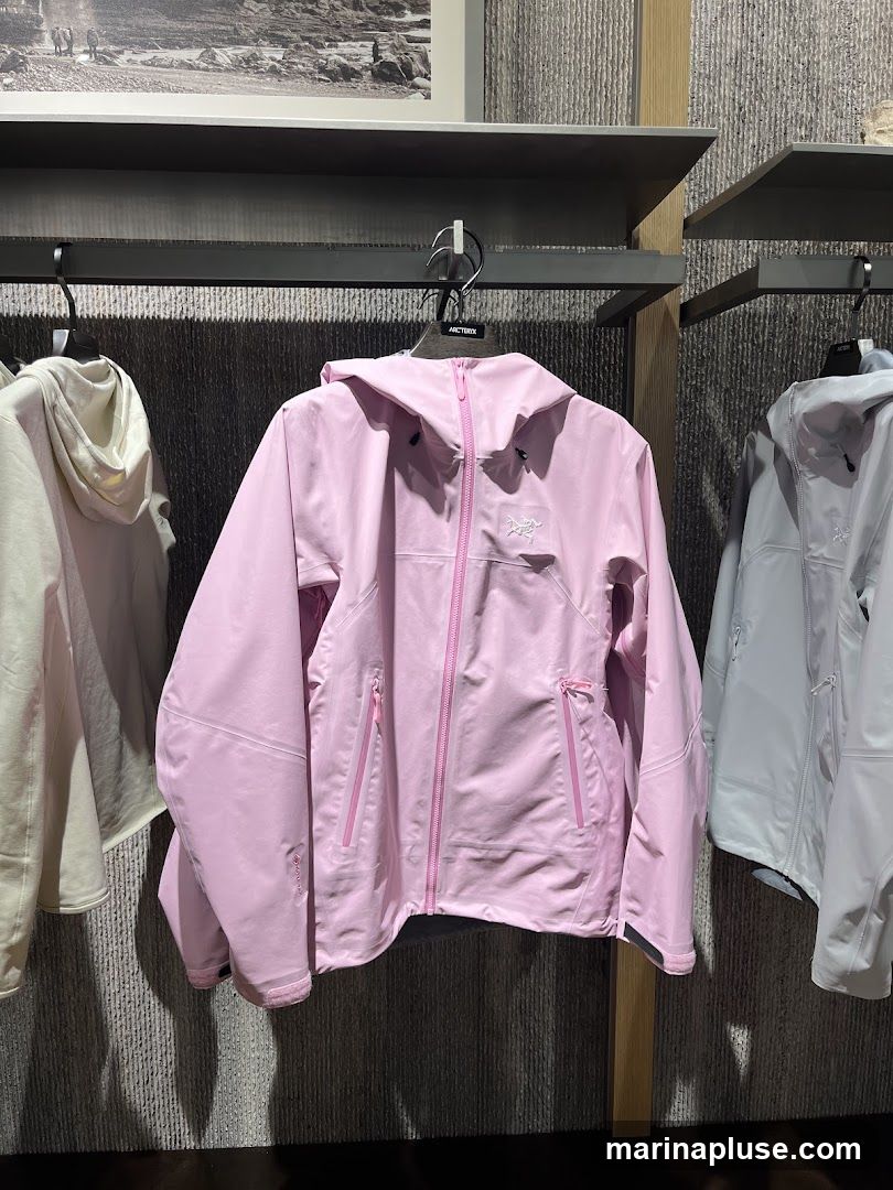 A pink Arc'teryx jacket is displayed on a hanger in a store. The jacket has a hood, a zipper closure, and zippered pockets. Another jacket in a light cream color is visible to the left, and a light grey jacket is to the right. A framed picture is on the wall above the display shelves.