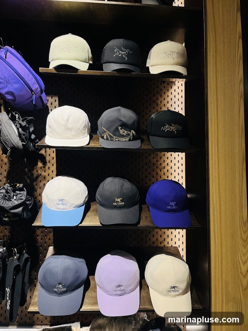 A display shelf filled with various hats and bags. The hats are arranged in rows, featuring different colors and designs, many with the Arc'teryx logo. There are also several bags, including a purple crossbody bag and black backpacks, visible on the left side of the display. The background is a wooden panel with peg holes.