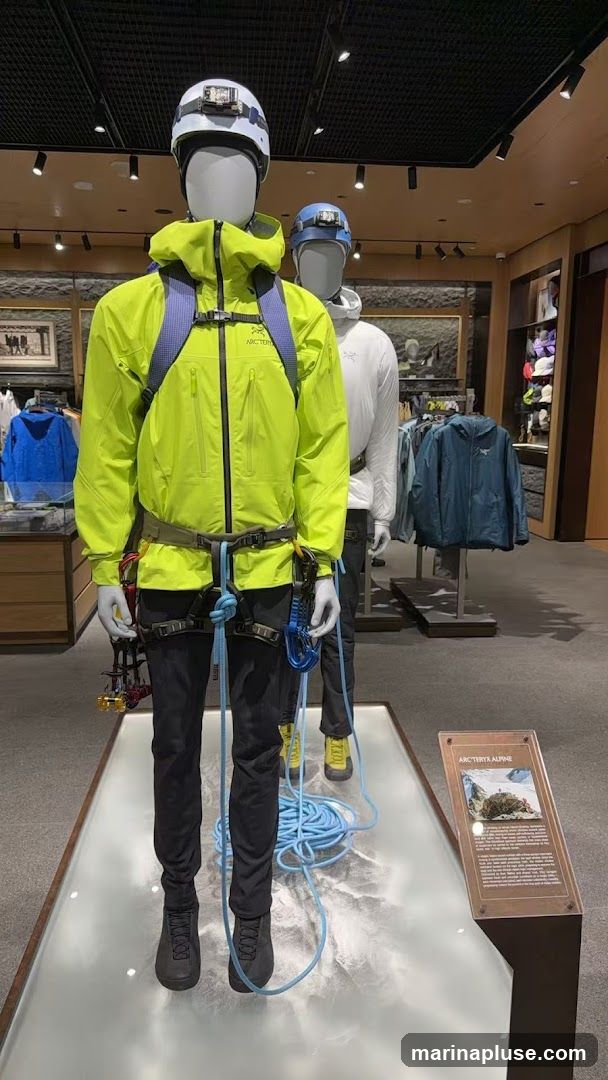 Mannequins dressed in outdoor climbing gear, including helmets with headlamps, brightly colored jackets, harnesses, and ropes, are displayed in a retail store. The mannequins are positioned on a platform with a map-like design. In the background, shelves display various outdoor apparel and accessories. A sign with the text "ARCTERYX ALPINE" and an image of a mountain scene is visible in the foreground.