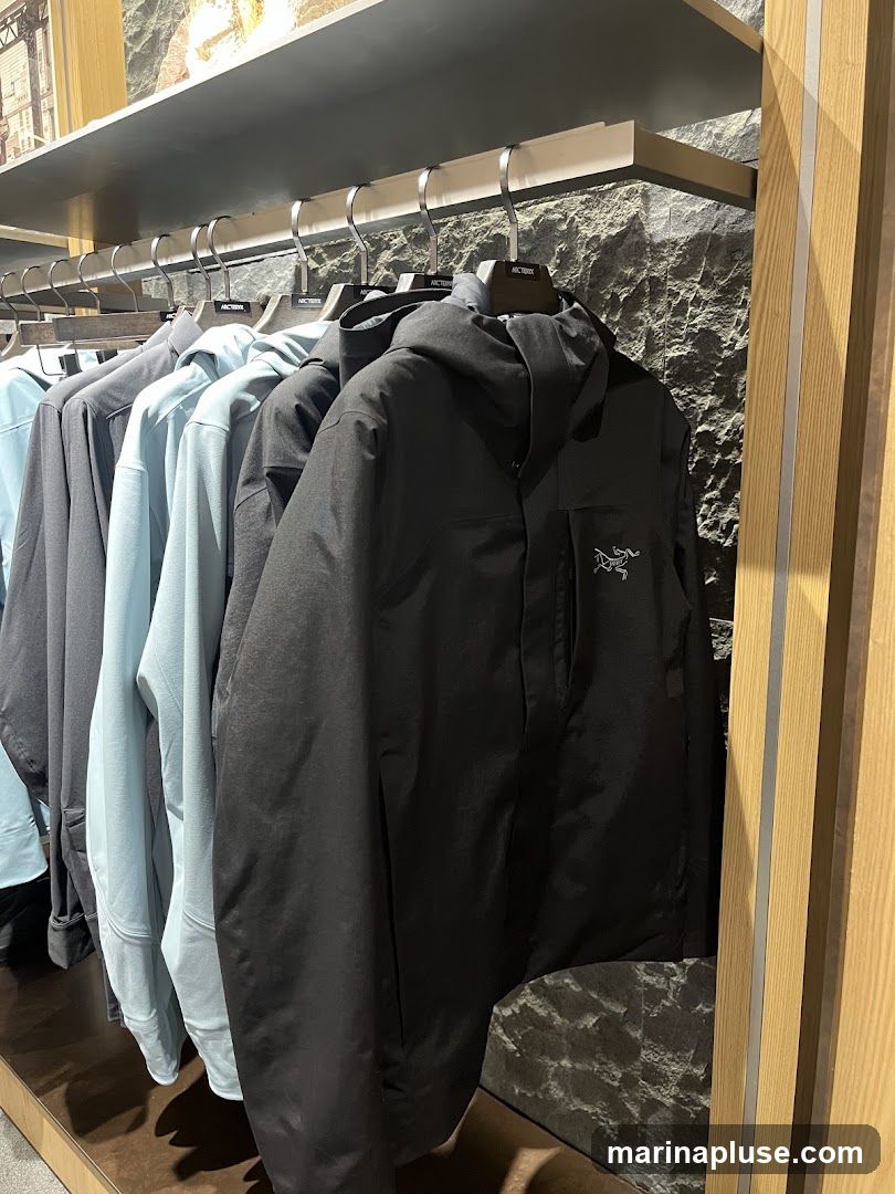 A rack of jackets and hoodies hangs on display in a store. The jackets are in various shades of gray and light blue. The foremost jacket is black and features a prominent Arc'teryx logo on the chest. The background has a stone-like texture and wooden accents.