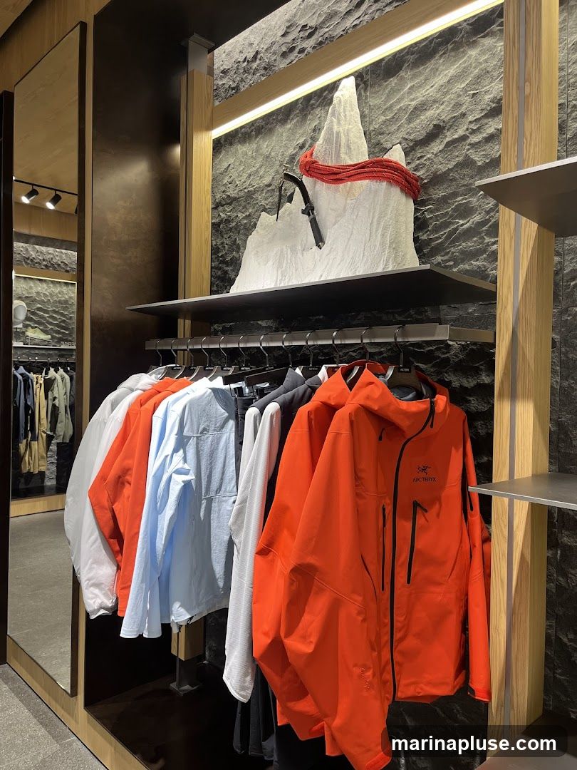 A retail store display featuring outdoor apparel. In the foreground, several jackets and shirts are hung on a rack, with a prominent orange jacket with the Arc'teryx logo visible. Behind the clothing, a decorative display of a white mountain peak with a red rope and an ice axe is mounted on a textured stone wall. The store interior includes wooden accents and mirrors reflecting more clothing racks.