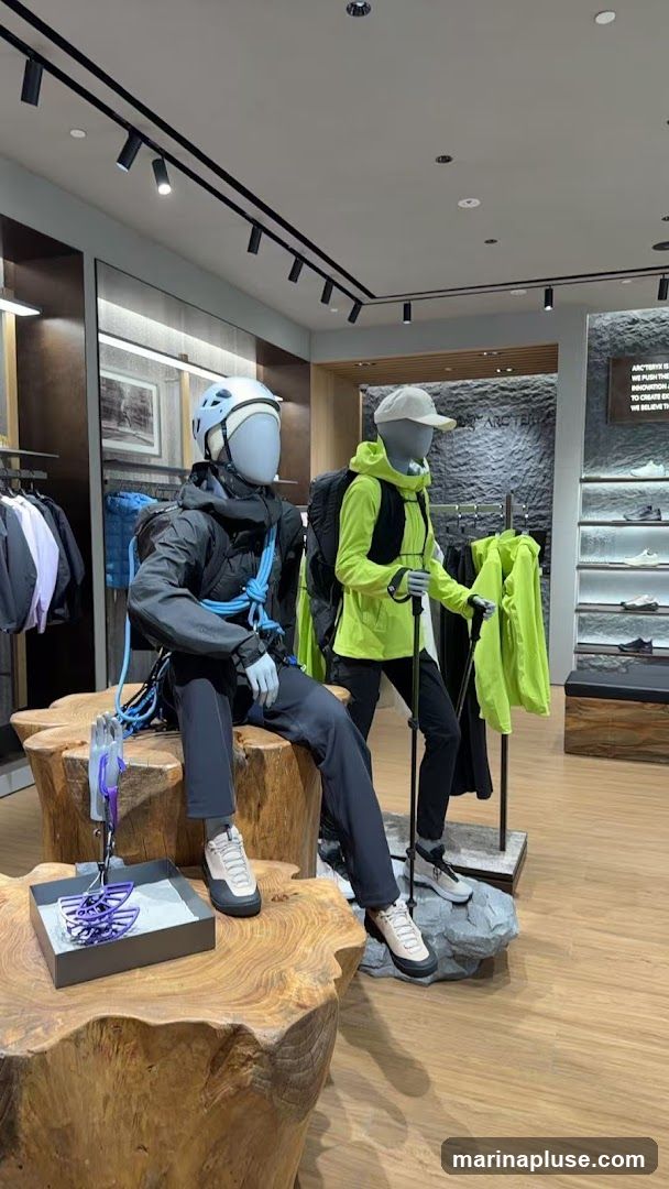 Mannequins dressed in outdoor gear, including helmets, jackets, backpacks, and hiking boots, are displayed in a store. One mannequin is seated on a wooden stump, while the other stands with hiking poles. A display box with purple climbing gear is on the stump. Shelving with shoes is visible in the background, along with clothing racks. A sign with text is mounted on the wall.