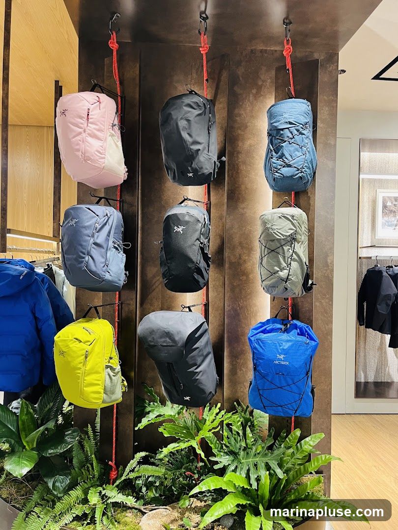 A retail display featuring nine backpacks of various colors and styles, including pink, black, blue, grey, and yellow. The backpacks are suspended by red ropes against a dark, textured wall. Lush green plants are arranged at the base of the display. In the background, clothing racks with jackets are visible.