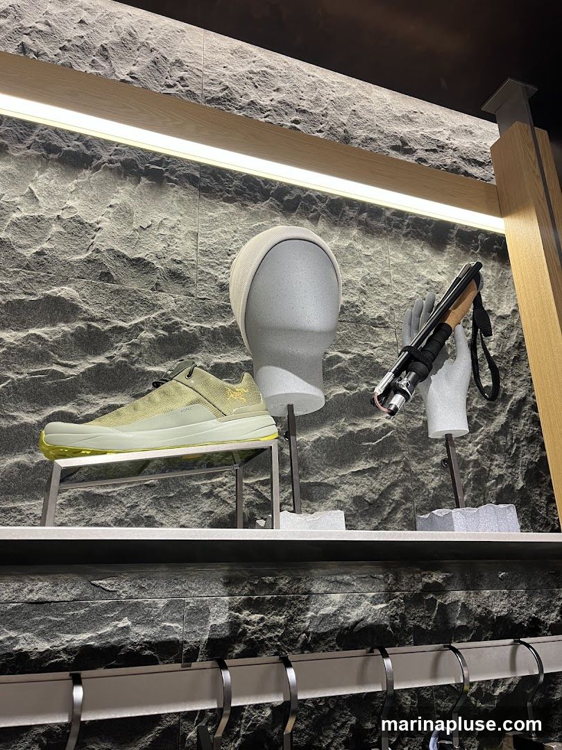 A display of outdoor gear including a light green athletic shoe, a grey beanie on a mannequin head, and a trekking pole held by a mannequin hand. The items are displayed against a textured grey stone-like wall with modern wooden accents and lighting. Below the display, hangers are visible on a shelf.